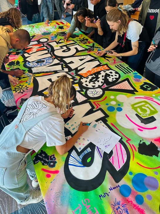 Groupe de professionnels en action lors d'une animation team building de graffiti&nbsp;: plusieurs personnes de tous &acirc;ges, &eacute;quip&eacute;es de bombes de peinture, travaillent ensemble sur un grand mur blanc ou gris dans un espace int&eacute;rieur type atelier. Certains sont en train de peindre, d'autres observent ou attendent leur tour, cr&eacute;ant une atmosph&egrave;re collaborative et ludique. L'ambiance doit &ecirc;tre dynamique, professionnelle et conviviale, montrant l'aspect social et participatif de l'activit&eacute; sans clich&eacute;s banals.