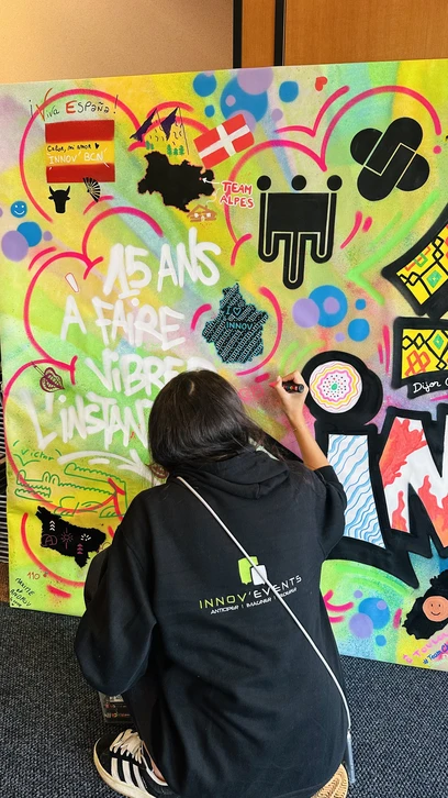 Photographie d'un groupe diversifi&eacute; de professionnels en tenue d&eacute;contract&eacute;e collaborant activement &agrave; la cr&eacute;ation d'une fresque murale color&eacute;e. Certains membres de l'&eacute;quipe tiennent des pinceaux et des bombes de peinture, appliquent des couleurs sur un mur blanc, tandis que d'autres observent avec engagement et participent au processus cr&eacute;atif. L'atmosph&egrave;re est positive, collaborative et &eacute;nergique, avec des sourires et une r&eacute;elle concentration artistique. Le d&eacute;cor peut &ecirc;tre un int&eacute;rieur professionnel (mur d'un loft ou d'un espace d'&eacute;v&eacute;nementiel) ou urbain. L'image doit capturer l'essence du live painting et de la fresque participative comme vecteur de coh&eacute;sion d'&eacute;quipe autour d'une cr&eacute;ation artistique commune.