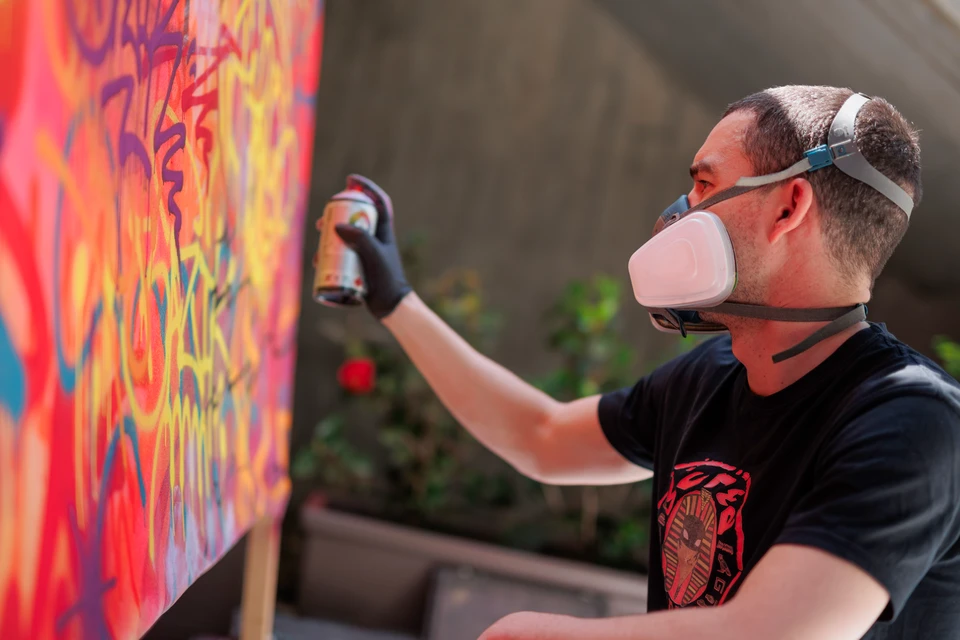 Photographie réaliste en couleur d'un artiste graffeur/peintre muraliste concentré en train de peindre une fresque murale. L'artiste, vu de profil ou trois-quarts, est en pleine création avec un pinceau ou spray, appliquant des détails avec précision sur un mur. Son expression faciale montre la concentration et l'implication. La fresque murale montre des éléments graphiques colorés en cours de réalisation. L'environnement est un vrai chantier ou espace de travail artistique, avec des équipements de peinture visibles. La photo capture l'engagement personnel et la maîtrise technique du professionnel en pleine action.