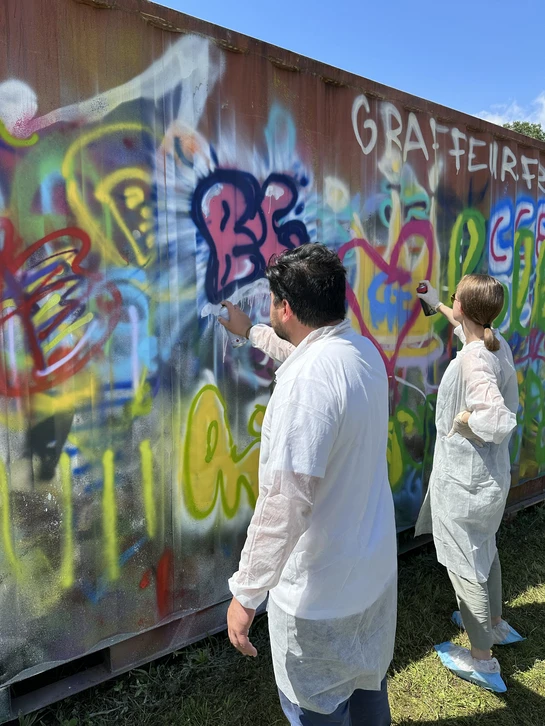 Photographie en couleur d'un groupe de 4 à 6 personnes en tenue décontractée travaillant ensemble sur une fresques de graffiti en cours de création sur un mur urbain. Au premier plan, une ou deux personnes manient les bombes de peinture aérosol avec concentration, en train de ajouter des touches colorées à l'œuvre collective. Les autres participants regardent, pointent des zones, discutent ou attendent leur tour, créant une atmosphère de collaboration engagée. L'œuvre en arrière-plan montrait des formes colorées et dynamiques typiques du style graffiti urbain. La lumière naturelle met en évidence l'interaction du groupe et le processus créatif en cours. L'image doit transmettre l'authenticitié, l'énergie créative et le véritable travail d'équipe dans un contexte de graffiti/street art professionnel.
