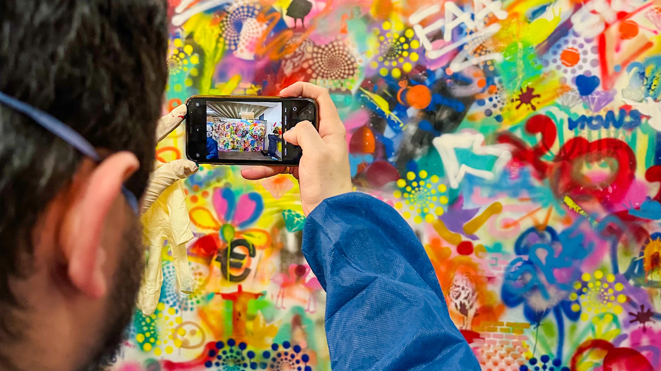 Groupe de professionnels d'une entreprise en tenue de travail (gilets, tabliers ou vêtements pratiques) en train de peindre collaborativement une fresque murale dans un espace intérieur ou extérieur. La scène montre des personnes de différents âges et profils tenant des pinceaux ou des outils de peinture, travaillant ensemble sur une paroi en cours de transformation. Des éléments graphiques colorés ou des motifs émergent sur le mur derrière eux, illustrant la création artistique en direct. L'atmosphère est dynamique, engagée, montrant clairement le processus de création collective et participative d'une œuvre murale.