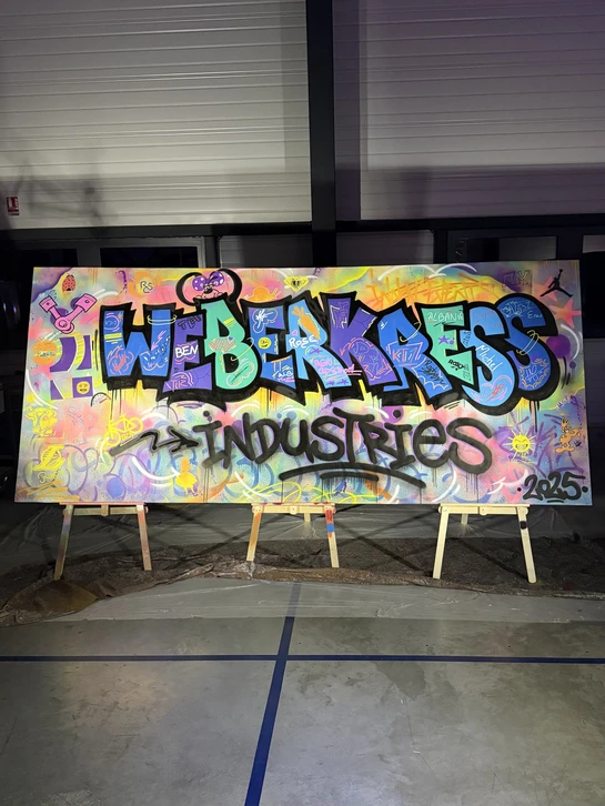 Photographie réaliste d'un atelier de fresque murale participative en cours d'exécution dans un espace professionnel (bureau ou salle). Plusieurs employés de l'entreprise, de diversités variées, sont debout ou en mouvement devant un grand mur blanc ou partiellement peint, munis de marqueurs, brosses ou bombes de peinture, collaborant activement à la création d'une composition colorée et artistique. L'atmosphère doit transmettre l'énergie, la collaboration et la créativité. La fresque en cours de création doit être visible et montrer des éléments graphiques modernes et professionnels. Aucun texte ni logo visible.