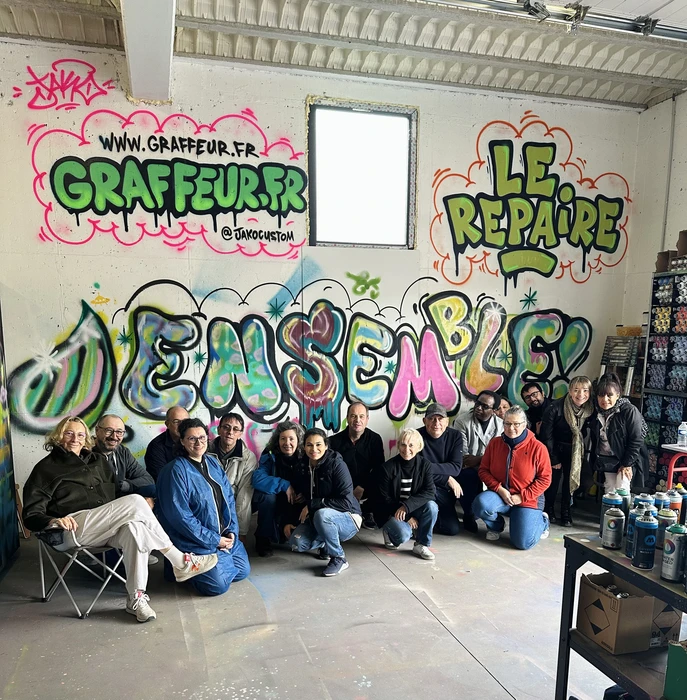 Photographie professionnelle d'une équipe de salariés d'entreprise en cours de création d'une fresque murale participative. La scène montre plusieurs personnes de profils et âges variés, certaines tenant des pinceaux ou des rouleaux, travaillant ensemble sur un grand mur intérieur coloré partiellement peint. Les participants affichent engagement et concentration. L'espace est lumineux, avec un rendu de fresque urbaine contemporaine aux formes géométriques ou abstraites. La photo capture l'atmosphère collaborative et créative d'un atelier de peinture murale en cours de réalisation, mettant l'accent sur la participation active du groupe.
