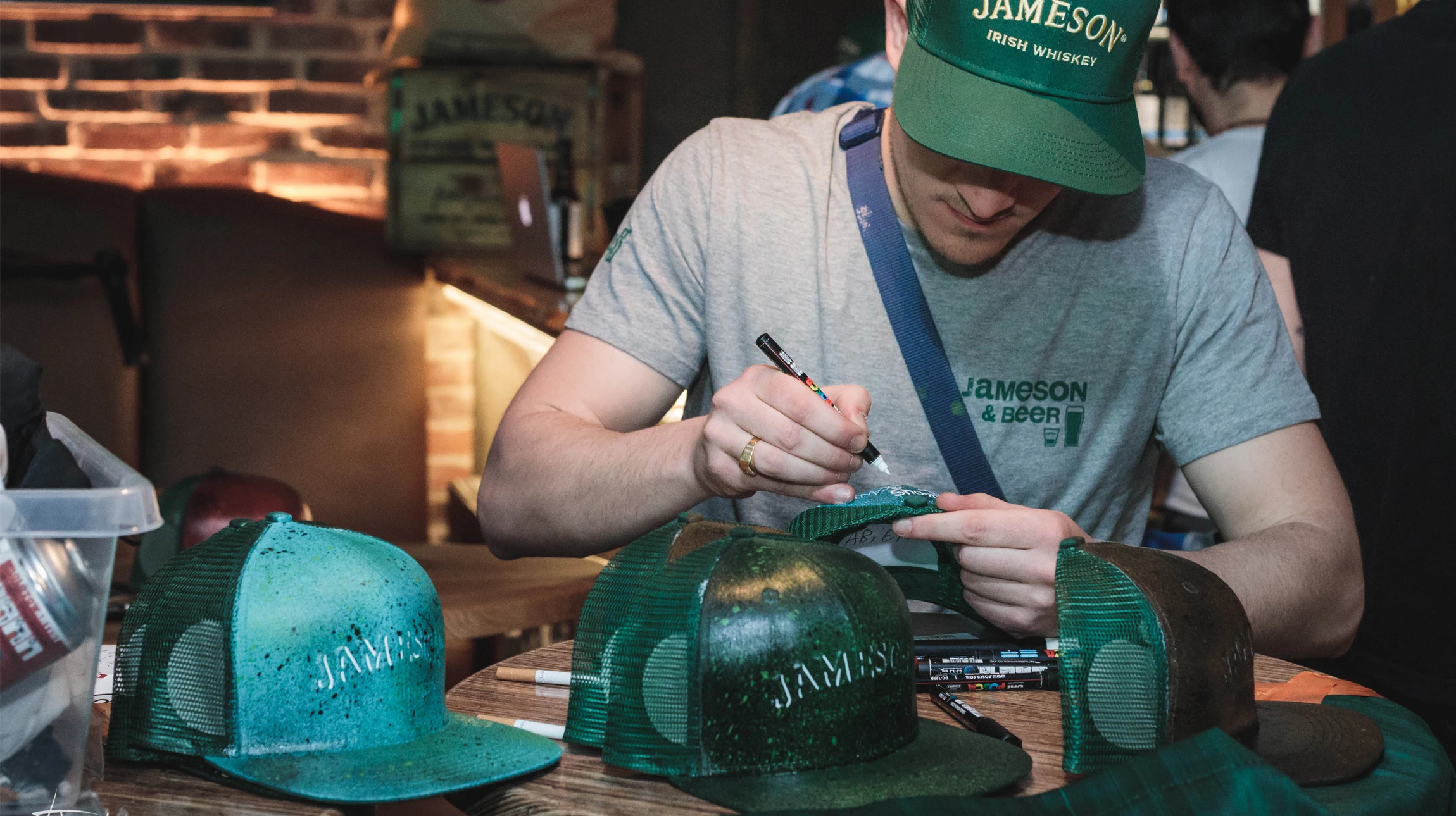 Scène en intérieur ou extérieur d'un événement professionnel montrant un artiste en train de personnaliser en direct un objet (casquette, t-shirt ou accessoire) avec des outils de décoration (marqueurs, peinture, aérographe) pendant qu'un groupe de collaborateurs ou de participants regarde, interagit ou attend son tour, créant une ambiance dynamique et fédératrice de team building. L'artiste doit être au cœur de l'action, concentré sur son création, dans un environnement qui évoque à la fois la créativité urbaine et le contexte professionnel de l'événement.