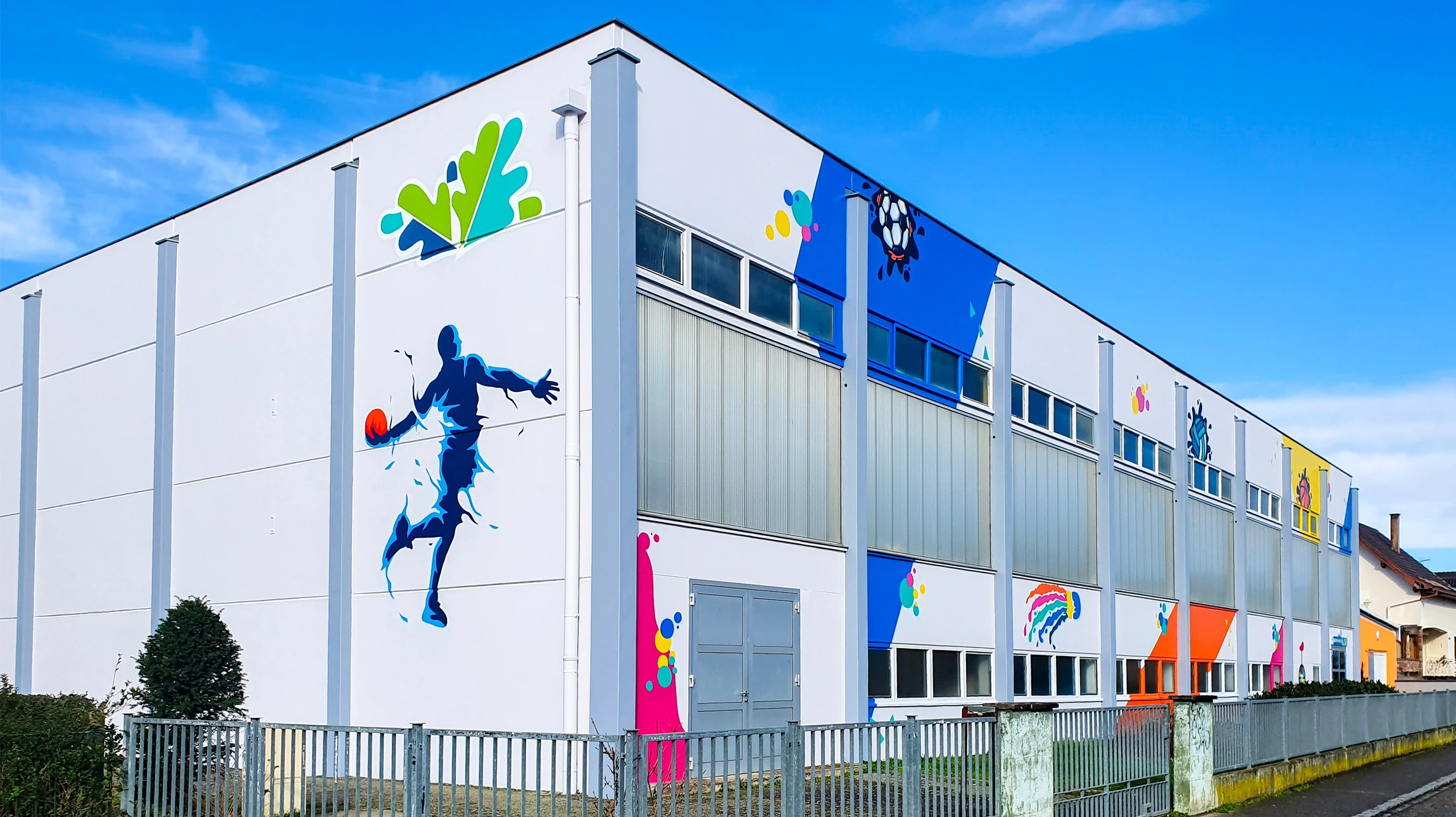 Photographie en prise de vue réelle montrant un artiste appliquant une fresque murale colorée sur la façade d'un bâtiment professionnel. L'artiste est en train de peindre au spray ou au pinceau, visible en action, portant une tenue de travail. La fresque en cours de réalisation doit être partielle, montrant des éléments graphiques contemporains et modernes, avec des couleurs vives et un design urbain. Le contexte architectural doit être clairement professionnel (façade de locaux commerciaux ou d'immeuble), en lumière naturelle, transmettant à la fois la maîtrise technique et la créativité artistique.