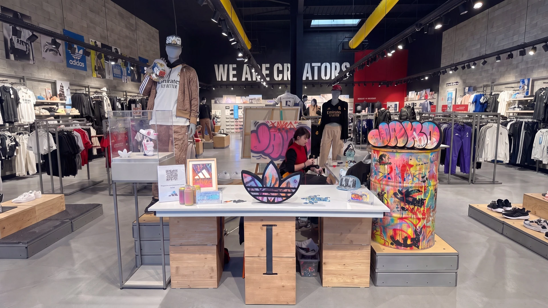 Artiste en atelier peignant à la main une casquette en coton blanc avec un marqueur acrylique coloré et un aérosol, reposant sur un plan de travail en bois brut. À proximité, une veste textile personnalisée avec des motifs graffiti multicolores séchant sur un cintre. Pinceaux, marqueurs Posca et bombe aérosol disposés sur le plan de travail. Ambiance d'atelier urbain, lumière naturelle, atmosphère créative et artisanale.
