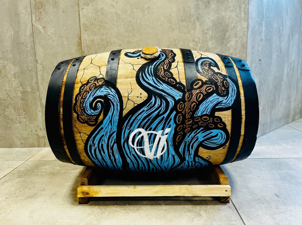 Casquette trucker noire posée sur surface en bois clair, customisée à la main avec un motif graphique coloré peint au marqueur acrylique ou à l'aérosol : formes géométriques, lettrages ou illustrations abstraites en rose vif, bleu cyan et jaune. Objet unique vue de trois-quarts face, éclairage naturel mettant en valeur les détails de la peinture artisanale.